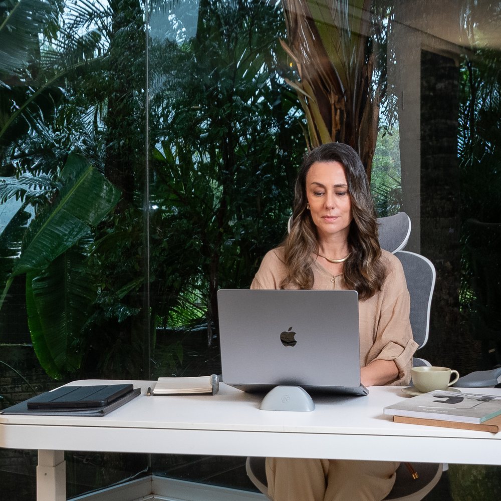 Mulher concentrada no laptop, de frente para a vidraça com vegetação